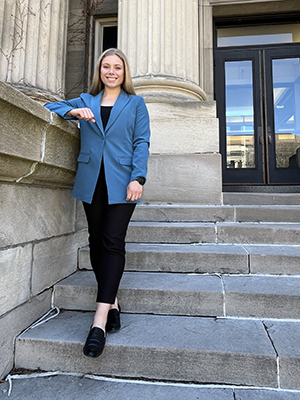 Paige Meyer stands on the steps of an East Bank campus brownstone building with tall sandstone columns behind her, her right elbow resting on a ledge