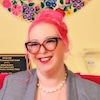 Stefani Goerlich sits in a pink chair at a pink desk. Stefani has pink hair in a bun, glasses, and wears a gray blazer over a yellow top.