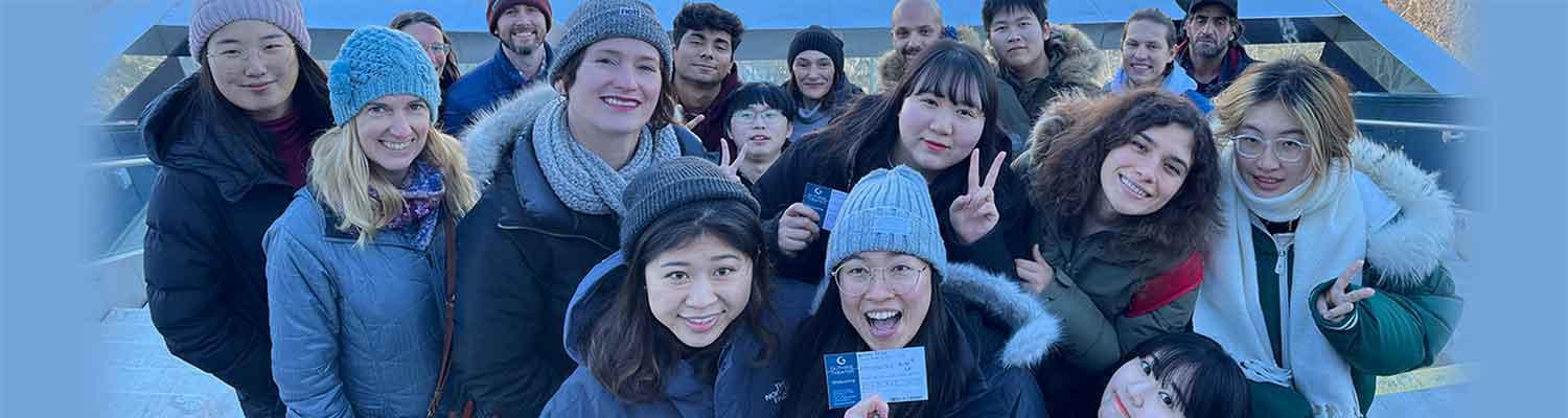 A group oof MELP students and instructors stand outside the Guthrie Theater's endless bridge in winter, smiling for the camera