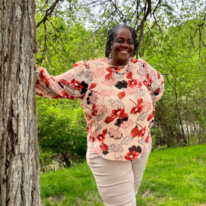 Shari Robinson stands with her right hand resting on the trunk of a large tree wearing an orange floral top