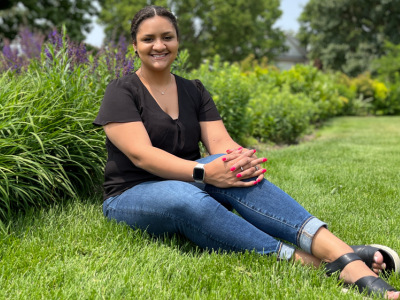 Aalayha Traub sits with legs outstretched and bent a little to the side and hands folded on her knees on the lawn on the St. Paul campus of the U of M with a perennial flower border behind her