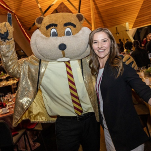 A woman stands arm-in-arm with Goldy Gopher, who is dressed in formal attire: a sparkling gold suit coat, gold shirt, and maroon and gold tie