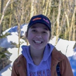 Gloria Kranenburg stands outside in a snowy forest ina  baseball cap, jacket, and sweatshirt.