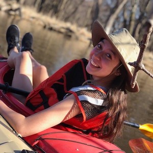 Gloria Kranenburg sits in a red kayak wearing a red life jacket and tan hat.