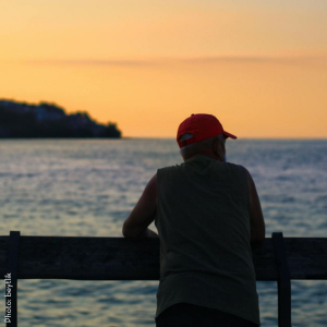 A man stands on a dock at sunset; his back is turned and he is watching the water