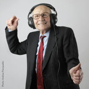 An older gentleman in a suit and red tie dances happily and with a grin on his face as he listens to music from headphones