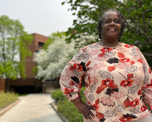 Shari Robinson stands with her right hand on hip gazing off into the distance, flowering trees and brick building in background
