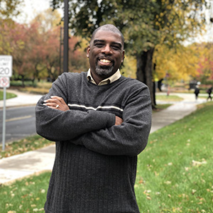 Black man wearing grey stiped sweater stands smiling with fall colored trees in background