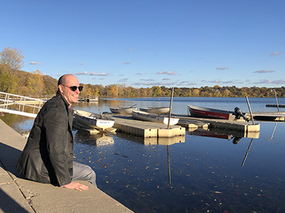 A bald white man dressed in blazer and jeans sits on a ledge overlooking Lake Harriet with boats and fall colored trees in background