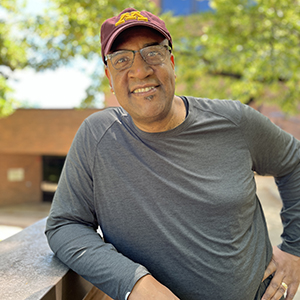 ITI Faculty Director Colin Miller, a Black man wearing a maroon and gold U of M ball cap and grey shirt and glasses, seen standing from the waist up, leaning against a brick wall and smiling