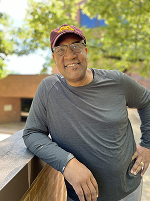 ITI Faculty Director Colin Miller, a Black man wearing a maroon and gold U of M ball cap and grey shirt and glasses, seen standing from the waist up, leaning against a brick walland smiling