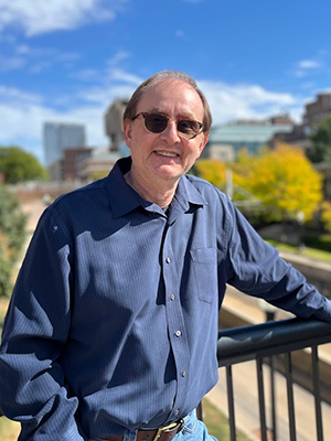 Carl Follstad leaning with elbow on black metal railing with U of M East Bank campus in background