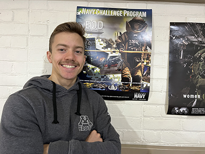 Owen Benson stands with arms crossed in front of poster for naval EOD program