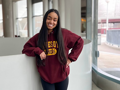 Soliana Ghidewon stands with right elbow on short white wall behind her and left hand on hip wearing a maroon and gold U of M hoodie and black leggings