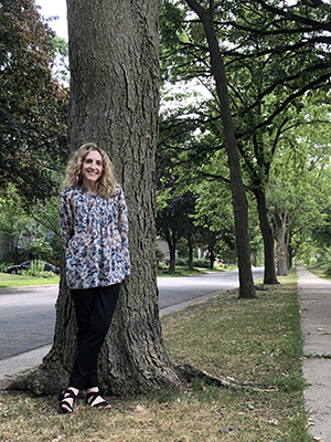 Blonde, wavy-haired, light-skinned woman leans against a tree, the first in a row of many trees lining a shady street