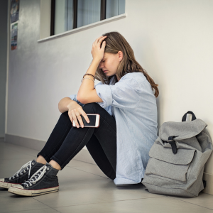a high school student holds her phone in one hand and  puts the other against her forehead in quiet distress as she sits against a wall in the school hallway with her backpack next to her