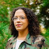 Lisa Marie Brimmer stands among green pine trees wearing a camouflage shirt and round glasses. They are Black and have long, dark wavy hair.
