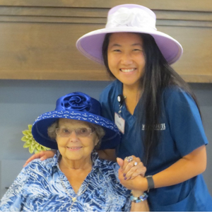 young woman healthcare worker wearing a large pink hat holds hands with an older woman resident who is wearing a large blue hat as both smile for the camera