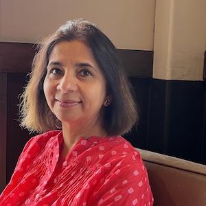 Neerja Singh, an Indian woman with shoulder length brown hair, wears a red shirt with small white flowers and sits in a chair..