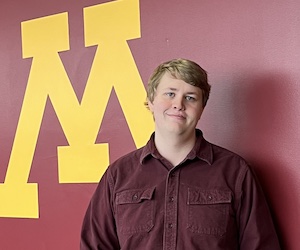 Noah Kincs in front of a gold block M painted on a maroon wall