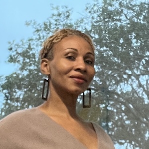 Shaun Telepak, a Black woman with short blond hair, stands in front of a large window with a tree in the background. She is wearing a tan dress and large silver rectangular earrings.