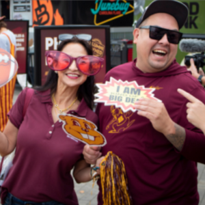 A woman wearing comically large plastic glasses and holding maroon and gold pom poms stands with a man who is laughing at someone who is off camera 