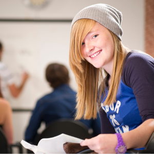 a high school student in a blue jersey and grey hat holds some papers and smiles for the camera while her teacher draws on a whiteboard in the background