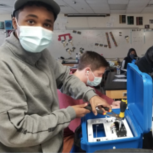 Young man wearing mask and hat works on a Solar Suitcase while another student works in the background