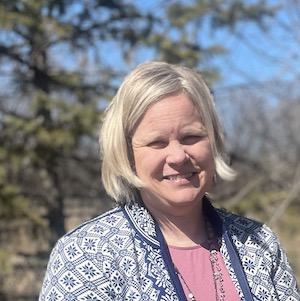 Teri Roath stands outside in the sun, looking at the camera, with an evergreen in the background. She wears a blue and white cardigan over a mauve shirt.
