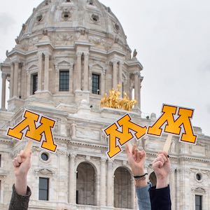Three gold M signs held up in front of capitol building
