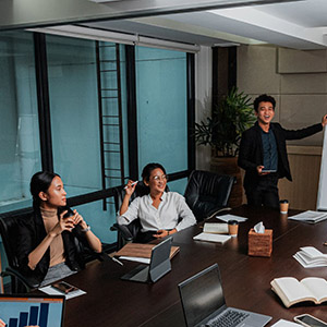 Young Asian man leads meeting at white board in front of table of other Asian professionals