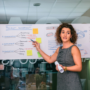 White curly haired woman in sleeveless dress stands before white board with project management terms and figures written on it