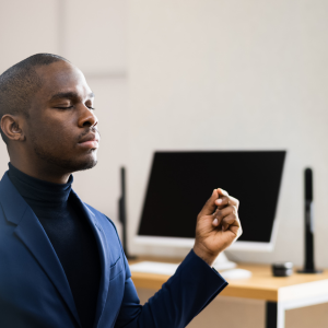 A Black man sits cross-legged on his desk with eyes closed and hands raised in mediative gesture