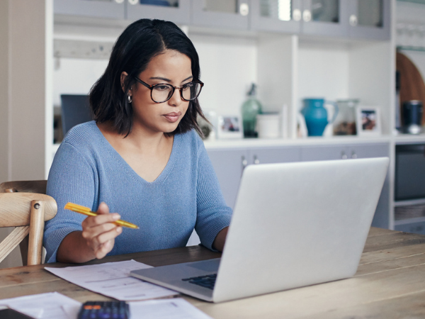 Hispanic woman in dark rimmed glasses looks intently at laptop holding pencil over a notepad with right hand