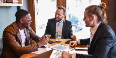 Three men sit at a table filled with papers while discussing business plans