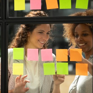 A white woman and light-skinned black woman discuss in front of a transparent wall covered in brightly colored sticky notes