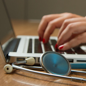 A woman types at a laptop with a stethoscope in the foreground