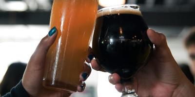 A Black woman's hand and a white man's hand raise glasses of beer in a toast. She's holding a hefeweisen and he's holding an imperial porter.