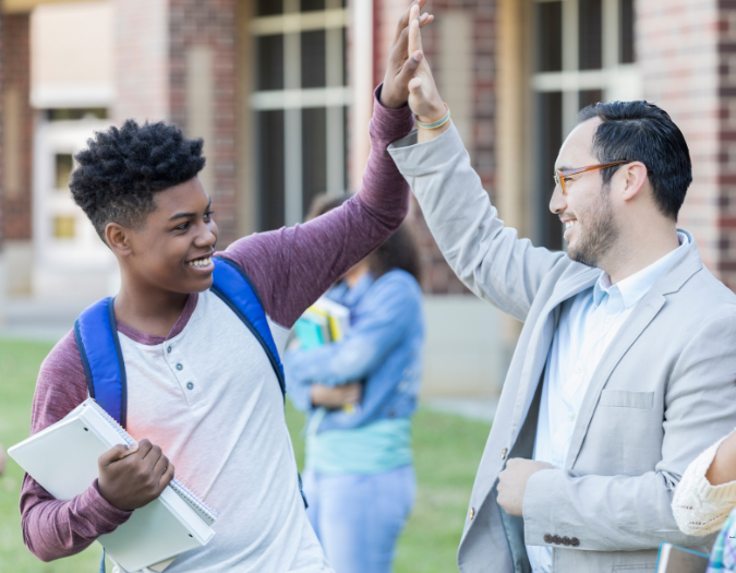 A high school administrator in a light blue suit coat high fives a smiling student on on the school lawn with other students looking on 