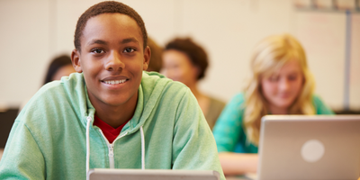 young student in green sweatshirt smiles at the camera while sitting at a table with his open computer in front of him 