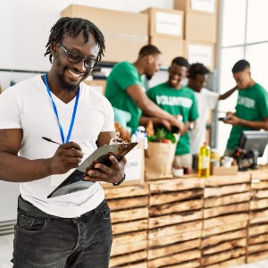 A Black man writes on a clipboard while a group of young Black men in volunteer shirts work in the background