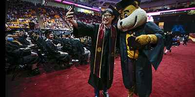 Male Asian student takes a selfie with Goldy, both wearing black cap and gown