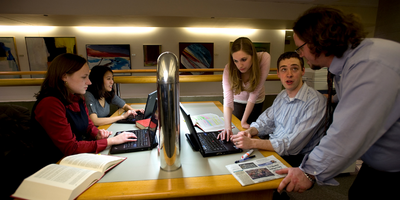 A group of students works around a library table with an instructor leaning over helping them