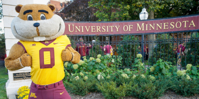 Goldy Gopher stands in front of an outdoor Twin Cities campus sign that reads University of Minnesota
