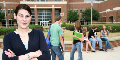 A high school administrator stands in front of the school she works at and looks at the camera with her arms folder. Students appear in the distance.