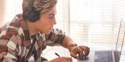 A high school students wearing a plaid short and headphones looks intently at a computer screen while also holding a pen