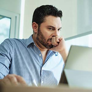 White man with scruffy beard and mustache leans on his fist with a grave look on his facing, looking at laptop