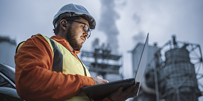 white bearded man with glasses, hard hat, and reflective vest studies laptop with industrial facility in background