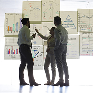 three professionals in office attire stand conferring in front of a large white board holding many charts and graphs