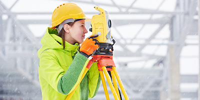 a white woman peers through a theodolite surveying a construction site
